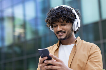 Close-up photo. Young smiling Indian male student standing outside wearing white headphones and holding phone. Calls, chats, listens to music
