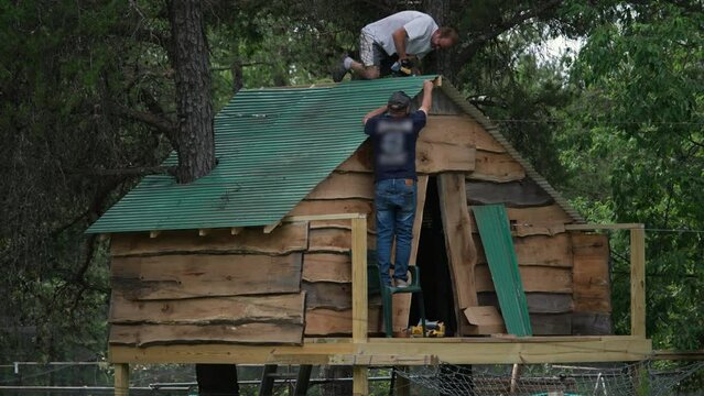 Men securing green sheets of metal onto roof of tree house build from scarp material around two trees.