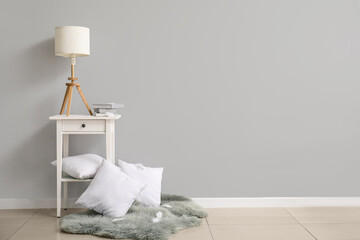 Bedside table with lamp and pillows near white wall