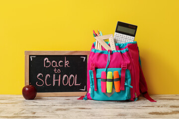 Colorful school backpack, stationery and blackboard with text BACK TO SCHOOL on wooden table near yellow wall