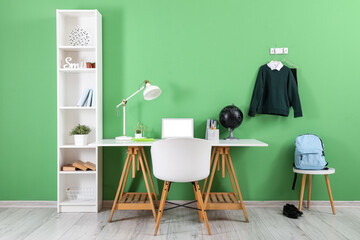 Workplace with desk, shelving unit, laptop computer, backpack and stylish school uniform hanging on green wall