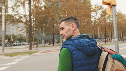 Mature man tourist wearing casual clothes, with backpack on his shoulder, looks around while standing on crosswalk