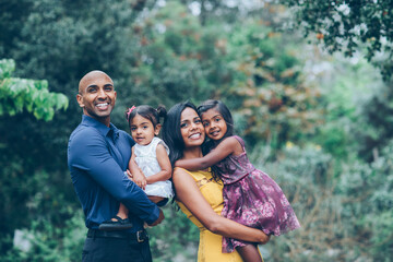 beautiful indian family standing and smiling holding their kids in the park with trees and greenery - Powered by Adobe