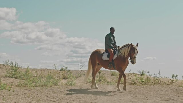Wide shot of young African American man riding chestnut horse in prairie on sunny day