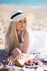 Beautiful blonde woman 26-28 year old wear straw hat lying on sand at beach over sea background close up. Summer vacation season.