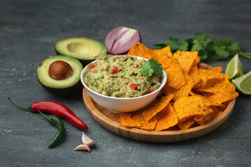 White bowl of traditional Mexican guacamole with nachos on grey concrete background. Tortilla chips with guacamole sauce dip and ingredients: avocado, cilantro, onion, hot pepper and lime