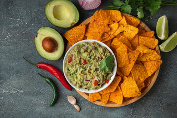 White bowl of traditional Mexican guacamole with nachos on grey concrete background. Tortilla chips with guacamole sauce dip and ingredients: avocado, cilantro, onion, hot pepper and lime. Flat lay