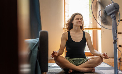 Young calm relaxed woman sitting at home on the floor in front of the fan in the lotus position