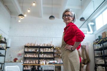Happy senior woman successful self-employed potter wearing apron behind table with pottery tools.