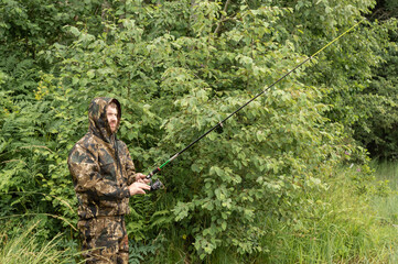 A young male fisherman stands on the shore of a lake. He has a fishing rod in his hand, looks intently at whether the fish are biting