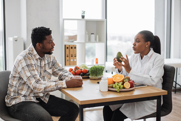 Handsome multiracial man in casual clothes visiting female dietitian in doctor's office of medical...