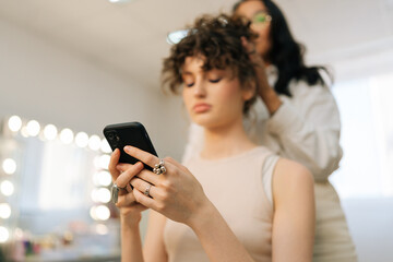 Closeup low-angle view of young woman client sitting in chair in beauty salon using phone while...