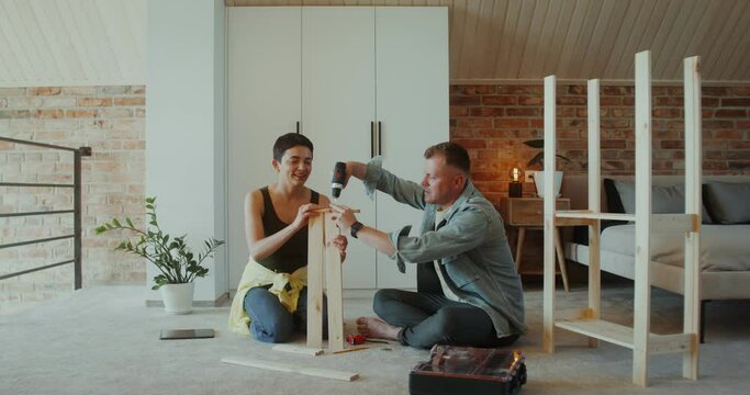 A man and a woman assemble a shelving at home, they use a screwdriver, talk and smile sitting on the floor