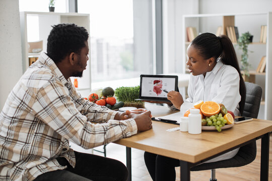 Confident Food Expert In Doctor's Office Pointing At GI Tract On Digital Screen While Talking To Male Patient On Midday. Multiethnic Woman Giving Advice On Healthy Diet For Digestive System Disorder.