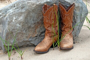 Brown western-style boots on sand by a large rock and green blades of grass 