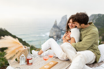 Coastal charms. Loving young couple embracing, drinking wine and enjoying picnic date with beautiful view on ocean shore
