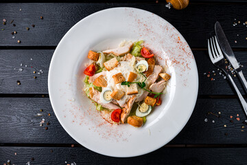 Caesar salad with chicken, parmesan cheese, eggs, lettuce, tomatoes, croutons on a dark table.