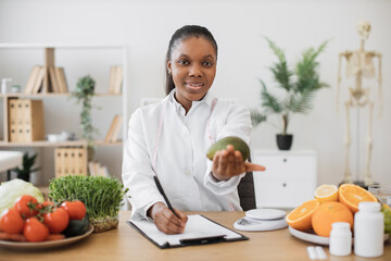 Portrait of smiling multicultural lady wearing white coat with avocado in hand posing at writing desk. Cheerful expert in food and nutrition selecting food for special diet in doctor's workplace.
