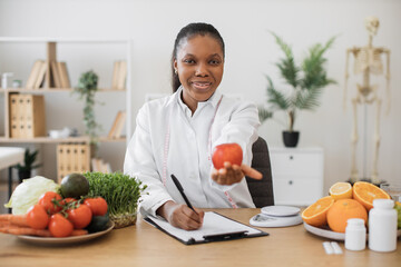 Happy multicultural female in white coat reaching out hand with fresh apple while working in dietitian's office. Efficient health professional encouraging to overall health and well-being via eating.