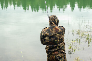 A young male fisherman stands on the shore of the lake with a fishing rod in his hands rear view