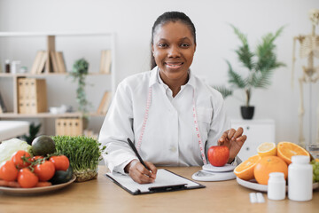 Focused multiracial woman in lab coat taking notes while assessing apple via dietary scale in...