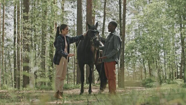 Full shot of young black man and caucasian woman standing together in forest on summer day, speaking and petting beautiful bay horse