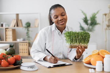 Positive female nutritionist with young sprouts in hand creating meal diet at writing desk in doctor's office. African american specialist with measuring tape recommending clients slimming foods.