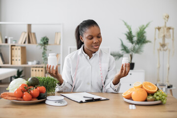 Focused female nutritionist holding two medicine bottles while working at office desk with products on it. Multiethnic health professional choosing food supplement for balancing diet of clients.