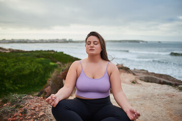 Fototapeta na wymiar Full figured woman meditating with closed eyes while sitting at gloomy wild beach