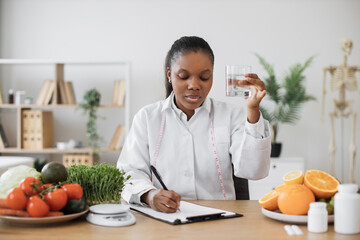 Thoughtful multiethnic lady with drink in hand making notes of diet changes while staying in...