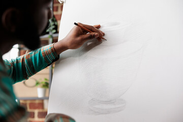 Student guy drawing with pencil on canvas. African American man artist staying focused while creating art in studio or workshop, sitting at easel sketching basic vase using pencil