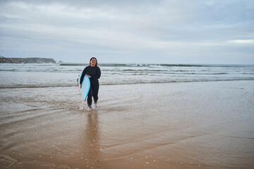 Positive surfer lady walking fas at the sea shore and feeling happy during cloudy day