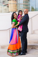 beautiful indian family with daughter girl hugging and smiling with a bindi and traditional sari dress in front of a building with staircase