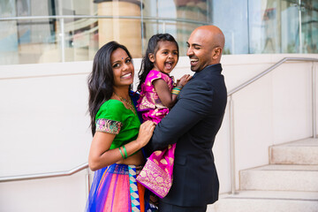 beautiful indian family with daughter girl hugging and smiling with a bindi and traditional sari dress in front of a building with staircase