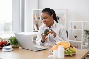 Charming adult lady in lab coat showing avocado to front camera of computer in professional office of clinic. Multicultural specialist in nutrition counseling patient online about benefits of fruit.