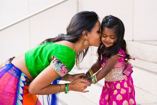 beautiful indian family mom mother with daughter girl holding hands kissing and smiling with a bindi and traditional sari dress in front of a building with staircase