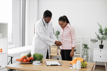 Multiethnic man in doctor's coat finding out waist length of female patient with measuring tape in...