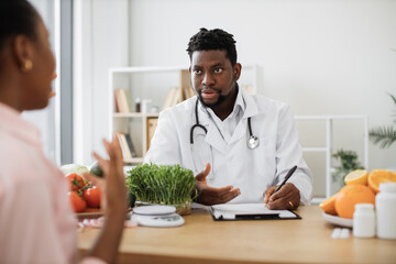 Fototapeta premium Confident man with stethoscope writing on paper clipboard during conversation with multiracial woman in office. Serious nutrition expert making notes of patient's medical history for proper meal plan.