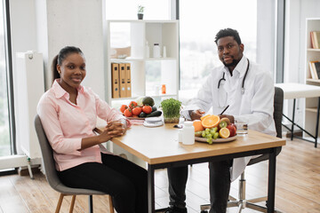 Full length portrait of multicultural people posing at office desk in doctor's workplace of medical center. Attractive woman in casual wear visiting professional dietitian on healthy eating issues.