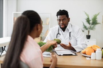 Focused african american man in white coat holding avocado while talking with female client in consulting room. Medical specialist in food encouraging to enriching eating with fruit full of nutrients.