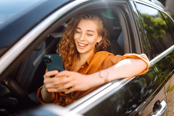 Happy woman in the car in the driver's seat looks at the smartphone. A young woman uses a mobile...