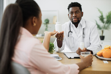 Obraz premium Handsome african american man in white coat giving pills bottle to charming woman in consulting room of hospital. Male nutrition professional improving overall health with dietary supplements.