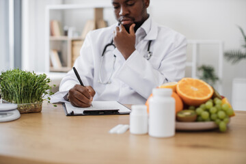 Expert in nutrition making special menu for patient and writing list of allowed products on clipboard paper during work time in hospital. African american man touching chin with hand and thinking.
