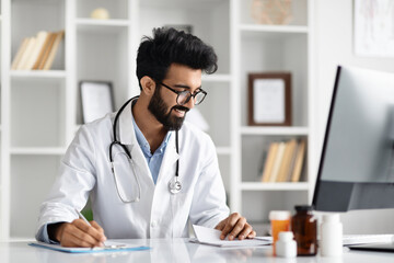Handsome indian man doctor using computer and writing notes - obrazy, fototapety, plakaty