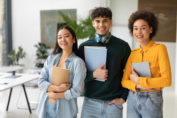 Happy three multiracial students posing in coworking space, young people with workbooks standing in classroom