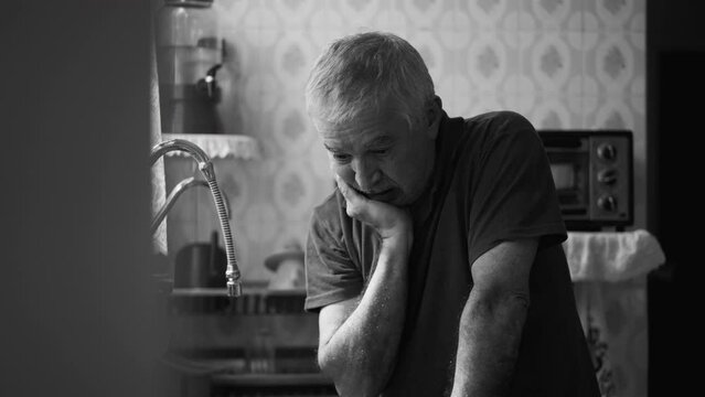 Senior man suffering from depression at home alone leaning by kitchen sink in dramatic black and white, monochromatic scene of person in despair succumbed by hardship