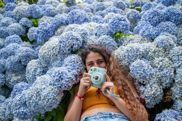 Beautiful young woman with long curly hair posing with a camera in her hands on a background of blue hydrangeas flowers. São Miguel island in the Azores