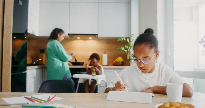 A girl draws with a stylus on a tablet while sitting at the kitchen table at home. Her family in the background is out of focus