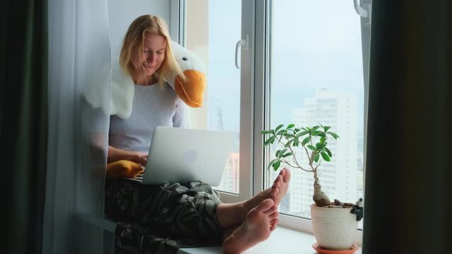 A happy barefoot woman using a laptop while sitting on a windowsill with a funny plush toy. Remote work, blogging, freelance.
