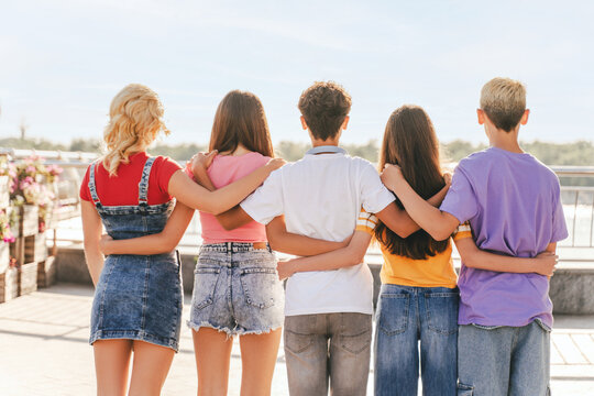 Back view group of friends, multiracial teenagers wearing colorful t shirts hugging each other looking away standing on the street. Friendship concept